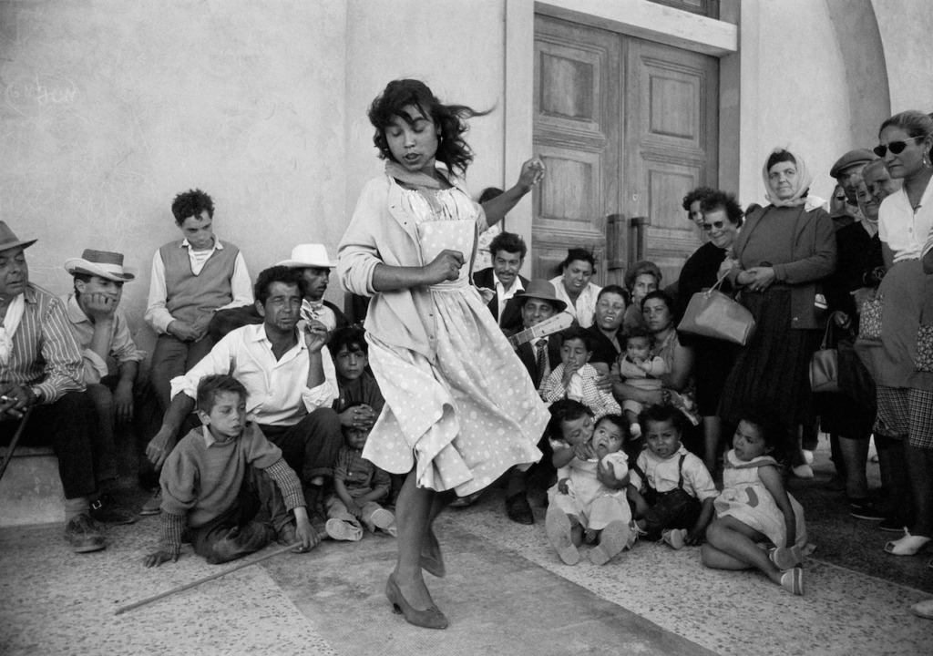 The picture shows a young woman in a white dress. She is dancing. In the background you can see women, men and children looking at her. Exhibition: Sabine Weiss. A Photographer's Life