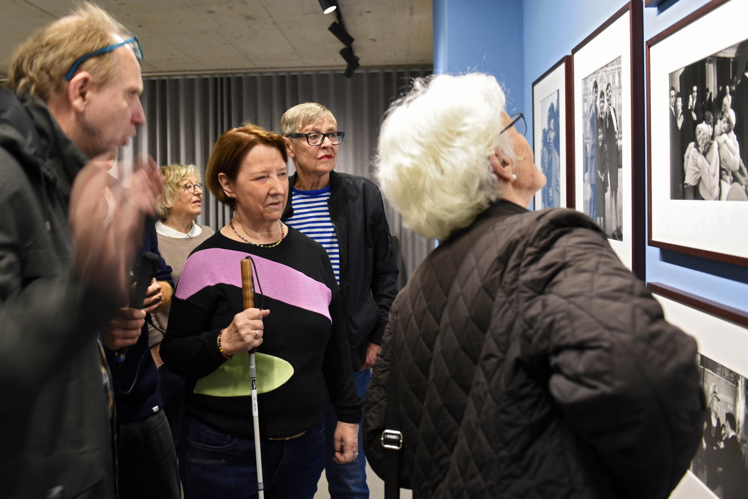 Eine Gruppe steht vor gerahmten Fotografien in einer Ausstellung. Eine Person mit Sehbeeinträchtigung führt die Gruppe durch die Ausstellung.